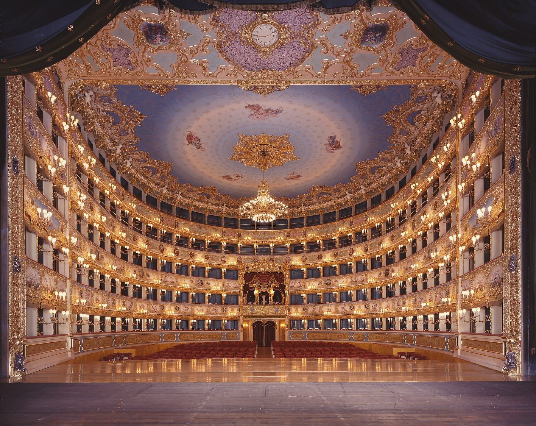 La sala del Teatro La Fenice vista dal palcoscenico: platea rossa, cinque ordini di palchi dorati illuminati e il soffitto azzurro con il grande lampadario