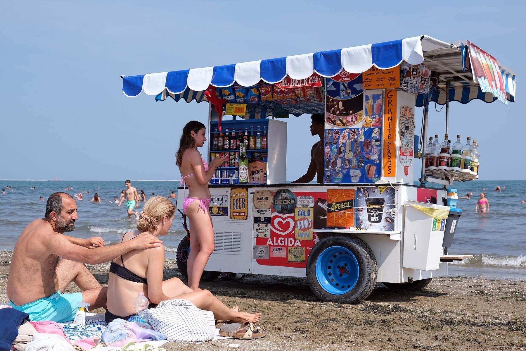 Spiaggia del Lido di Venezia con bagnanti sulla sabbia e un carretto di bibite e gelati con tendone a righe bianche e blu, il mare sullo sfondo
